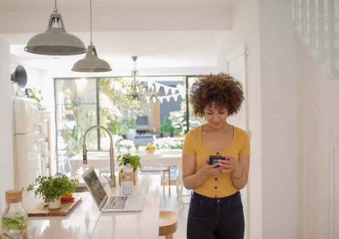 Young woman using smart phone and computer to check health savings account standing in sunny kitchen.