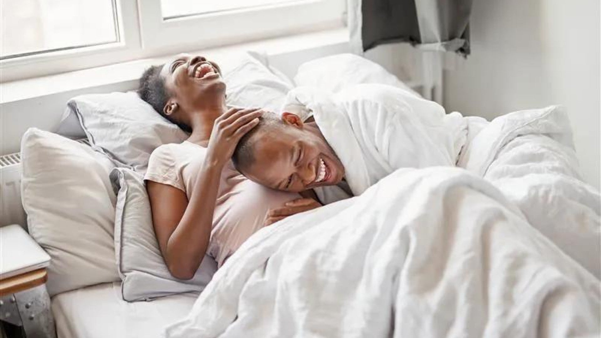 Young couple laughing together while relaxing in bed with white bedding, sharing a joyful morning moment