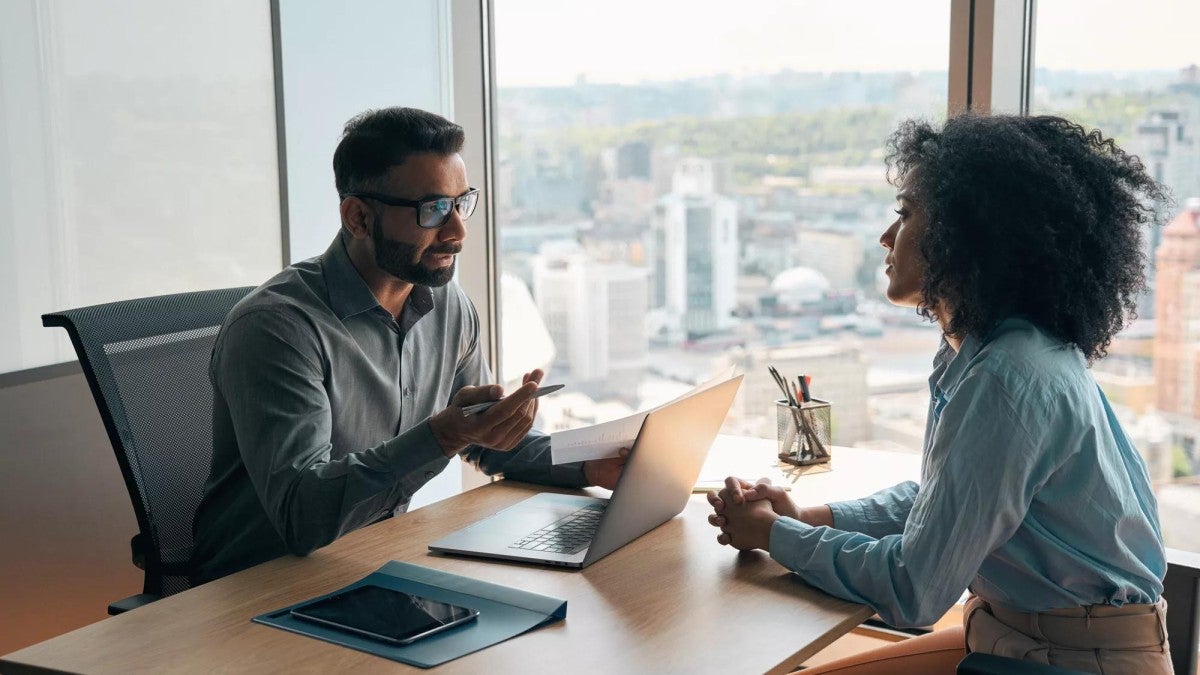 A man and a woman talk while seated at a desk in an office.