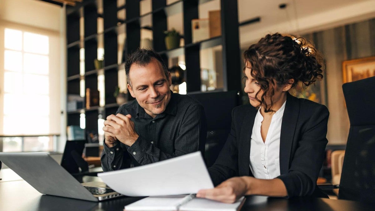 A man and a woman talk while seated at a desk in an office.