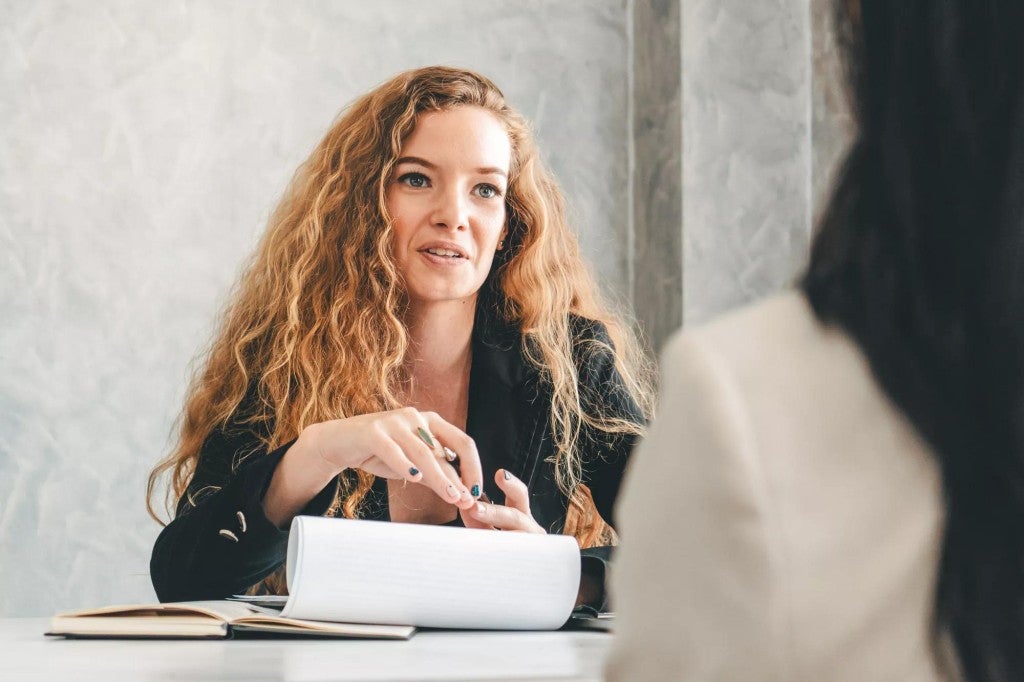 Woman in a meeting room speaking to another person with their back to the camera.