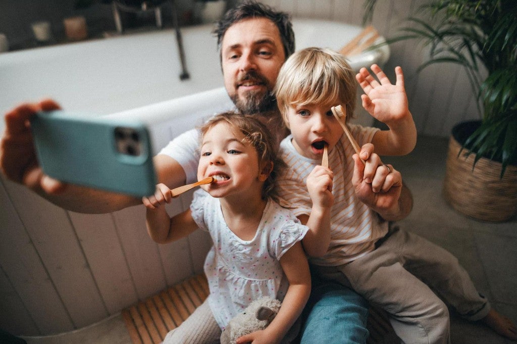 A man taking a selfie with his two young children while they are brushing their teeth.