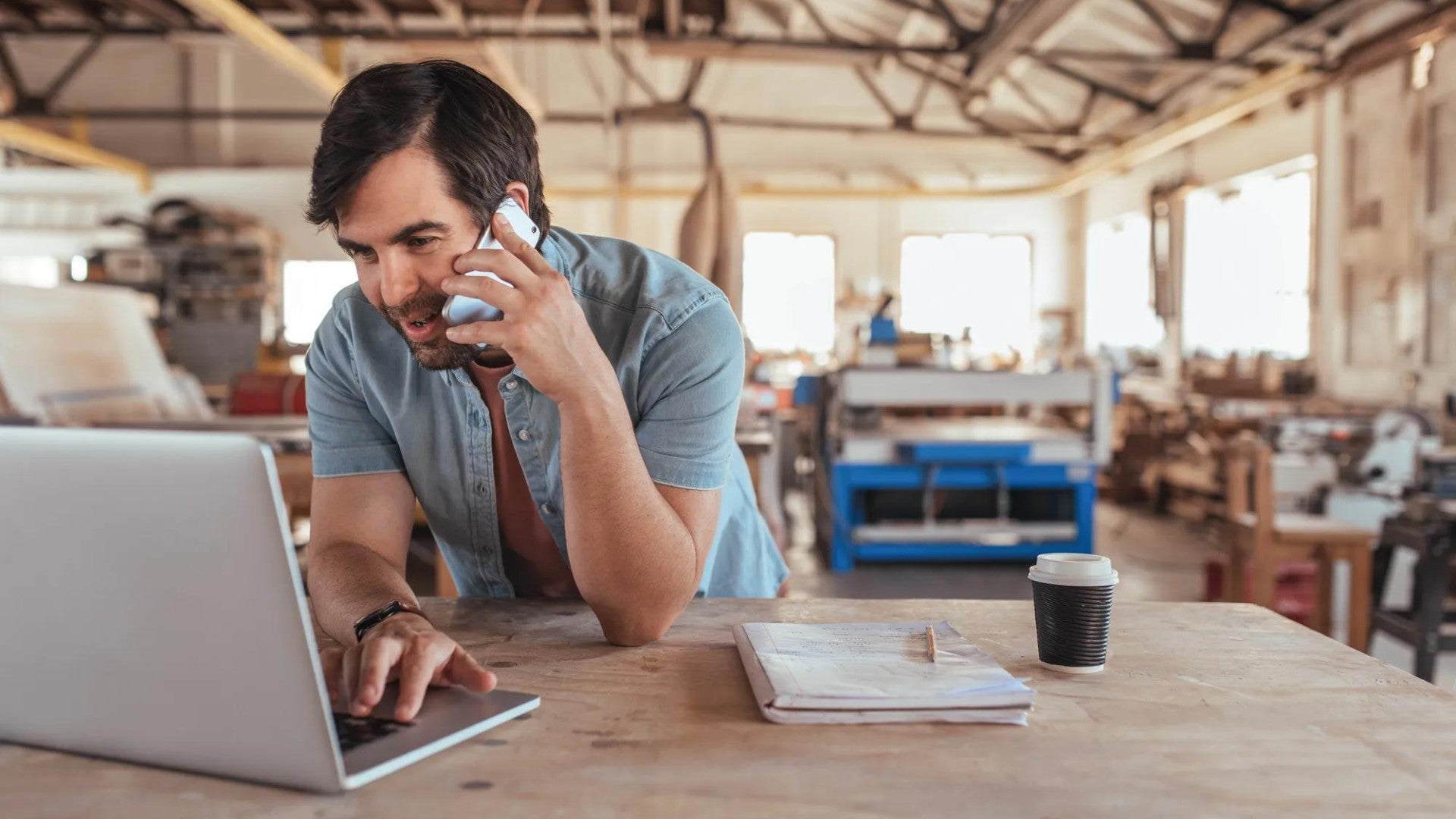 Man in shop area of small business talking on phone and looking at computer.